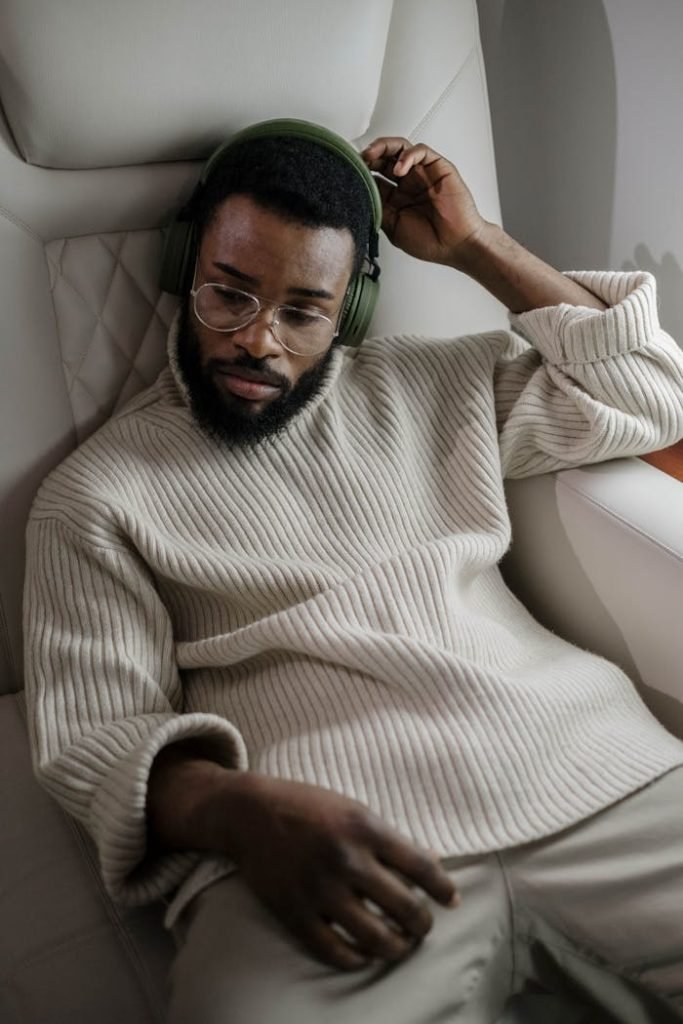 Bearded man with glasses enjoying music on a flight, wearing cozy sweater.