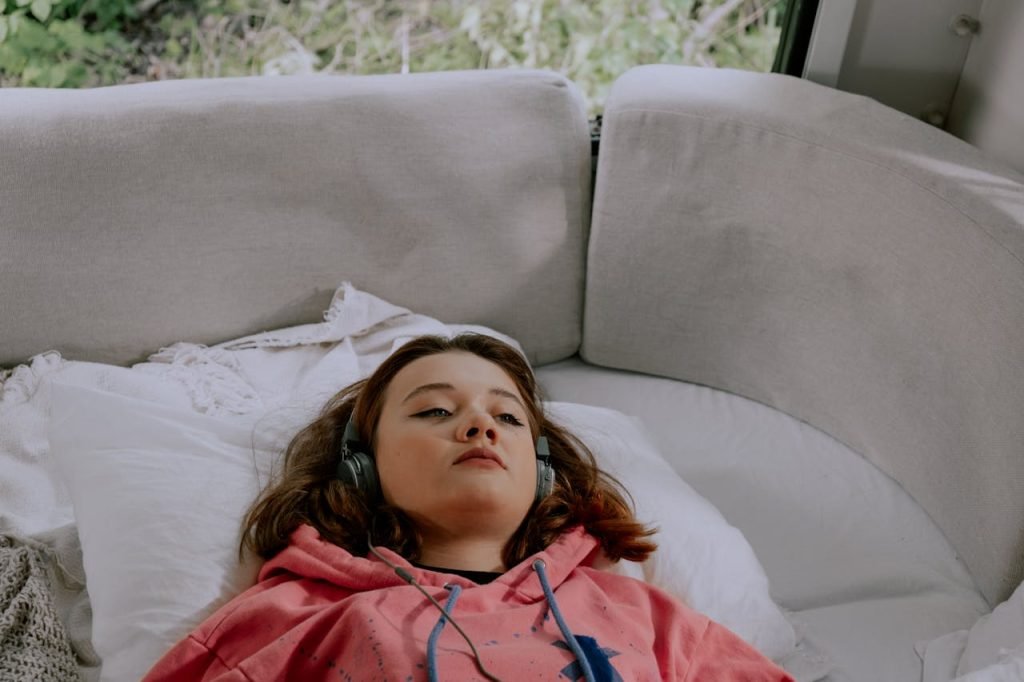 Woman in a pink hoodie lying on a sofa with headphones, enjoying music.