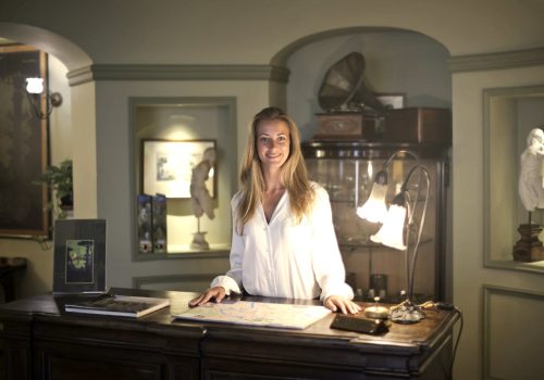 hospitable-woman-standing-behind-front-desk-3771087 Caucasian woman smiling behind hotel reception desk, creating a warm and welcoming atmosphere.