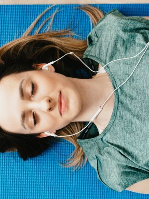 tranquil-woman-resting-on-yoga-mat-in-earphones-at-home-4498187 Young woman meditating with closed eyes on a yoga mat, listening to music.